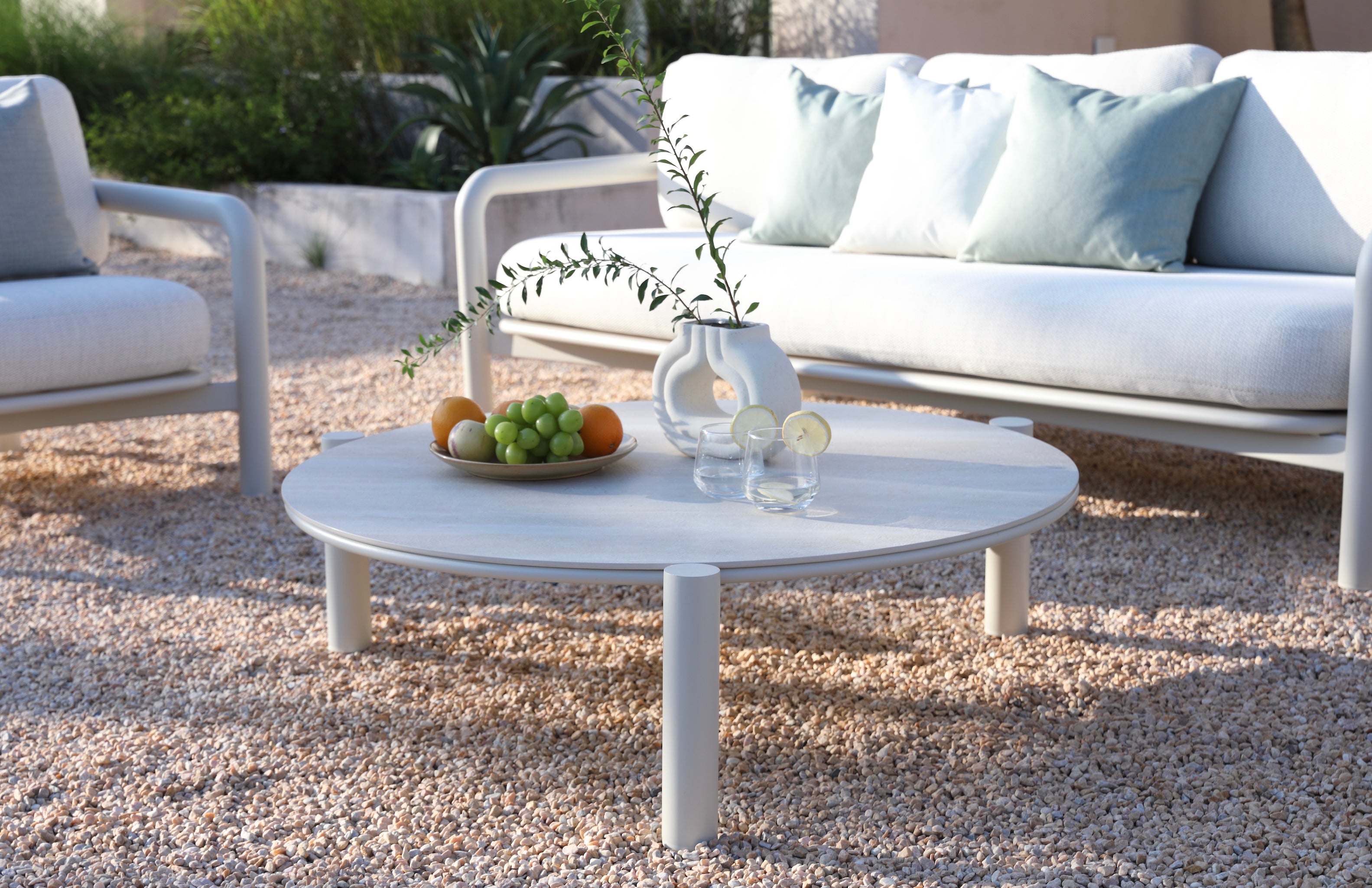 Table basse ronde avec structure cylindrique en aluminium blanc, plateau céramique lisse, entourée de sièges au tissu.